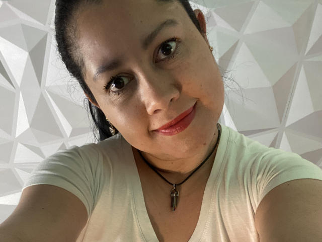 Close-up of MarianaSmitt with light brown skin, dark brown eyes, and black hair in a ponytail. She's smiling with red lips, wearing a white V-neck shirt and a black necklace with a small cross. Her eyebrows are dark and slightly arched. The light is even, highlighting her facial features. The geometric pattern in the background is subtle and white. She's looking slightly downward at the camera. Her skin has a few small blemishes. The image is focused and clear.