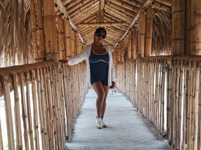 JessicaFiore with brown skin, short dark hair, small tits, wearing a blue dress, white cardigan, and white sneakers, standing in a bamboo-covered walkway, smiling, arms relaxed.