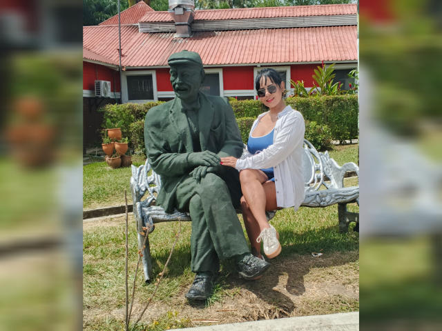 JessicaFiore met grote borsten, zittend op een bank naast een groene standbeeld van een oude man met een hoed, gekleed in een witte labjas en een blauw badpak..