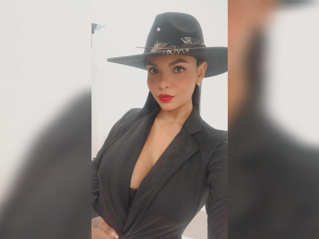 JessicaFiore met rode lippenstift, een zwarte hoed en een zwarte blouse met een diepe V-hals, waarbij haar borsten zichtbaar zijn, staat voor een spiegel, met een reflectie aan de rechterkant..