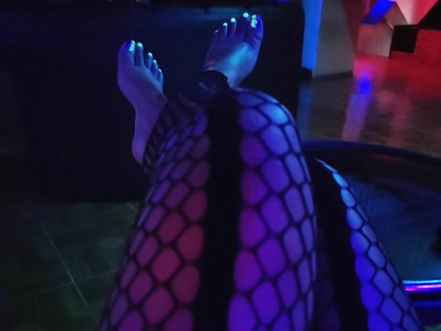 DolceCate's feet in black fishnet stockings, with glowing green toenails, crossed at the ankles, illuminated by blue and purple lights. Her legs are bent, showing the pattern.