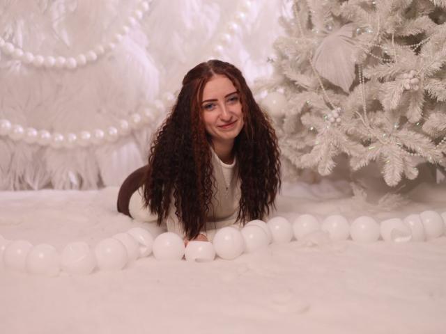 SandraPrafer with curly brown hair, white long-sleeve shirt, lying in a white ball pit, smiling, next to a white Christmas tree, surrounded by white pearls.