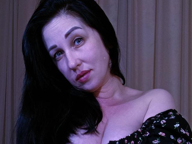 JillSilvies with pale skin, dark hair, and blue eyes, wearing an off-shoulder floral top, showing her tits, has a slight pout, and looks directly at the camera.