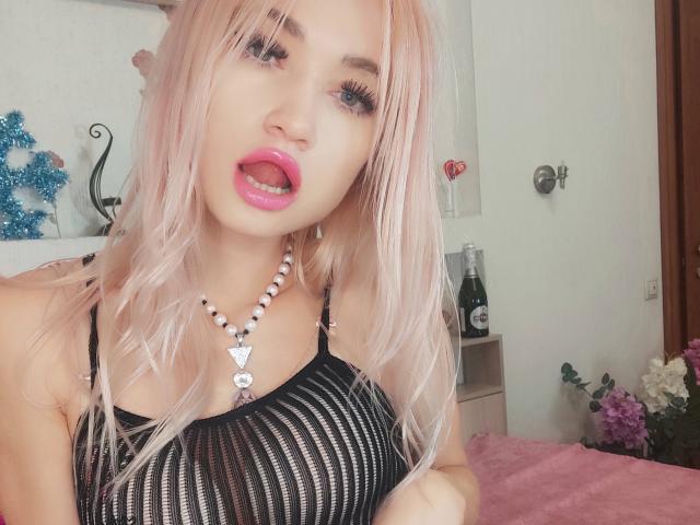 AlinaFit with pink hair, black mesh top, pink lips, tongue out, bead necklace, showing her tits, blue eyes, long eyelashes, in a bedroom.