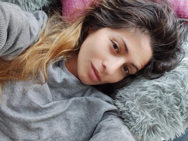 SkylerSamuel has wavy brown hair with blonde ends, wearing a gray hoodie, lying on fluffy gray and pink pillows, looking relaxed with a subtle, calm expression.