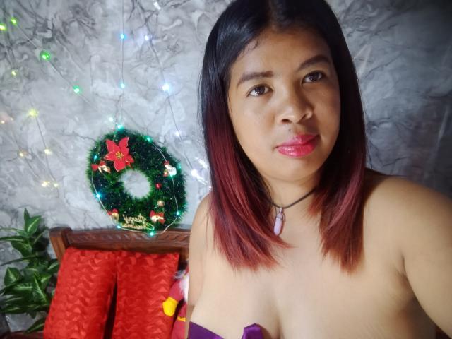 MariahSquirting69 with red-brown hair, pink lipstick, necklace, and purple strapless top, showing her tits, wearing a Christmas wreath necklace, sitting in front of red-patterned chair.
