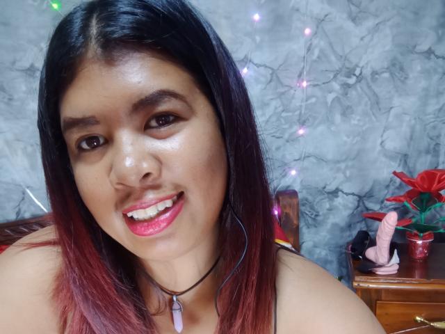 MariahSquirting69 tiene el cabello castaño oscuro con puntas rojas, lleva lápiz labial rosa, está sonriendo y tiene brillo rosa en los dientes, y un collar negro con un colgante morado..
