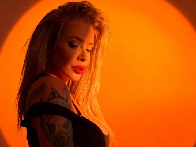 GlamSadie with long blonde hair, red lipstick, and a tattoo on her right shoulder, wearing a black top, illuminated by an orange circular light. Her eyes are closed.
