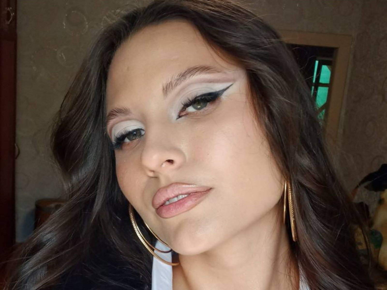 LeslieSvilti's face is centered in the image, showing a close-up. She has long brown hair, slightly wavy, and is looking to the left with a subtle, confident expression. She's wearing bold, white eyeshadow with black winged eyeliner, and pinkish-nude lipstick. Her skin is smooth, and she's wearing large gold hoop earrings. The lighting is natural, highlighting her makeup and facial features. Her lips are slightly parted. The background is blurred, with a window and patterned wallpaper. The image is a clear, focused portrait.
