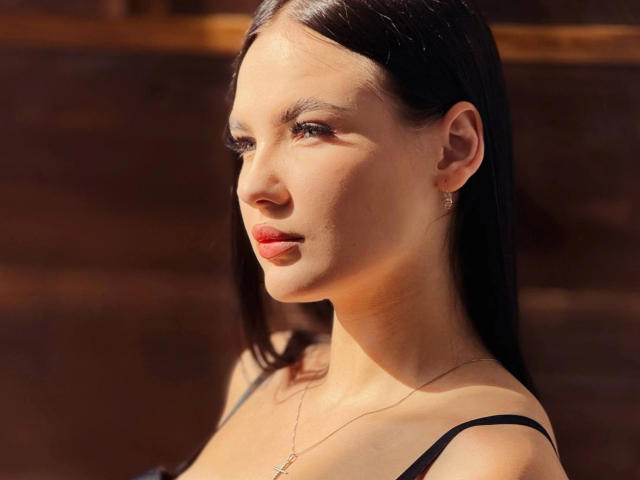 MilanaBeautty-hot is a close-up portrait of a woman with fair skin, straight black hair, and subtle makeup. She has a slender face with defined cheekbones, small earrings, and a delicate necklace. She wears a black strap top, looking to the right with a slight, confident smile. The light is warm, highlighting her smooth skin and the shadows in her hair. Her eyes are focused and her lips are slightly parted. The image has a soft, natural light that enhances her facial features.