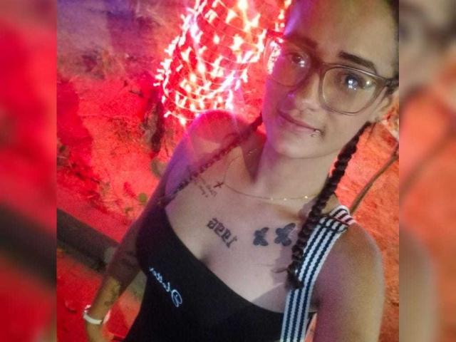 CattaleyaEvans with braided hair, glasses, black tank top showing tits and "beed" tattoo, and small nose piercing, in red-lit setting.