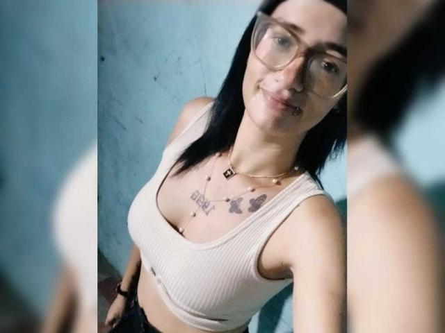 CattaleyaEvans mit Brille, schwarzem Haar, weißem Rippstrick-Tanktop, kleinen Brüsten, Schmetterlingstätowierungen, lächelnd, goldener Halskette, blauer Wand, Spiegelreflexion..