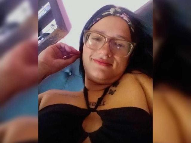 Cattaleya Evans con gli occhiali, top nero senza spalline che mostra il seno, foulard floreale, piercing al naso, tatuaggio sulla spalla destra, sorridente, che tiene un telefono, divano blu, primo piano sfocato..