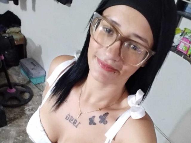 CattaleyaEvans com lenço preto na cabeça, óculos, blusa branca, mostrando os seios, tatuagens "BBR" e de borboleta, piercing no nariz, sorriso discreto, em um selfie casual..