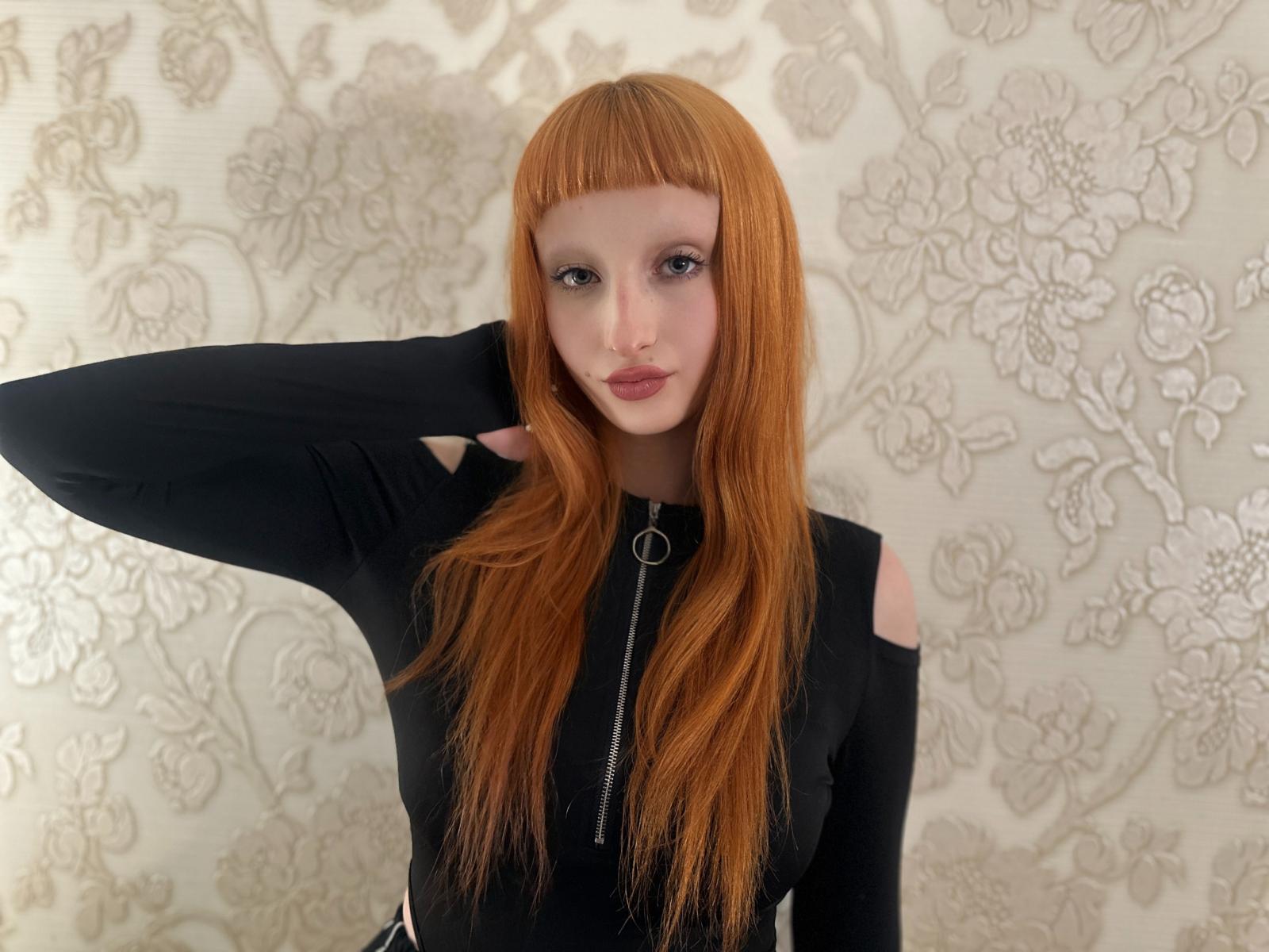 JaneWatson with long red hair, wearing a black zip-up top with cutouts, hand in hair, looking at camera, pale skin, subtle makeup, pink lips, floral patterned wallpaper.