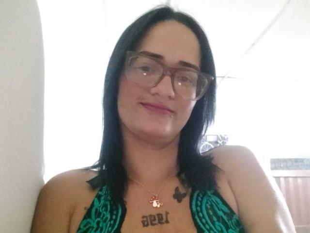 CattaleyaEvans with black hair, glasses, and green patterned dress, showing small tits and tattoo above her chest that reads "Deri." She's smiling with a slight nose piercing.