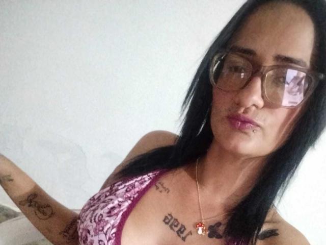 CattaleyaEvans with black hair, glasses, purple patterned bikini top, tattoos on chest and arm, lip piercing, gold necklace, white wall background, slightly pouty expression.