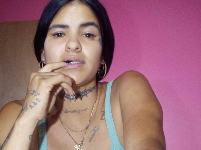 BeautifulTattoo with short black hair, brown skin, tattoos on neck and hand, wearing a green tank top, gold necklace, and hoop earrings, biting her finger with a slight smile.