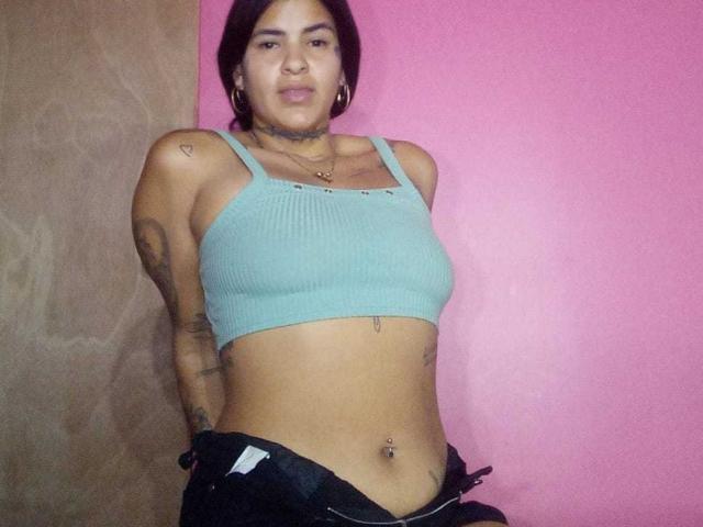 A photograph of BeautifulTattoo standing against a pink wall. She has a medium build, tan skin, and short dark hair. She's wearing a light blue ribbed crop tank top, exposing her midriff and small breasts, with black pants partially pulled down. She has multiple tattoos on her arms, a choker necklace, and a navel piercing. Her expression is neutral. The background is a pink wall on the right and a brown wall on the left. The image is candid with a casual, unrefined style.