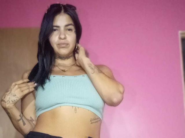 A photograph of BeautifulTattoo with long black hair, light brown skin, and tattoos on her arms and stomach. She wears a light blue ribbed crop top and black pants. Her right hand touches her hair while her left hand rests near her neck. She has a subtle smile and wears a gold necklace. The pink background is plain. The image is well-lit with soft shadows.