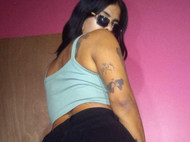 BeautifulTattoo with dark hair, wearing sunglasses, a light green tank top, and black pants, showing tattoos on her arm and back, with a pink wall and wooden door.
