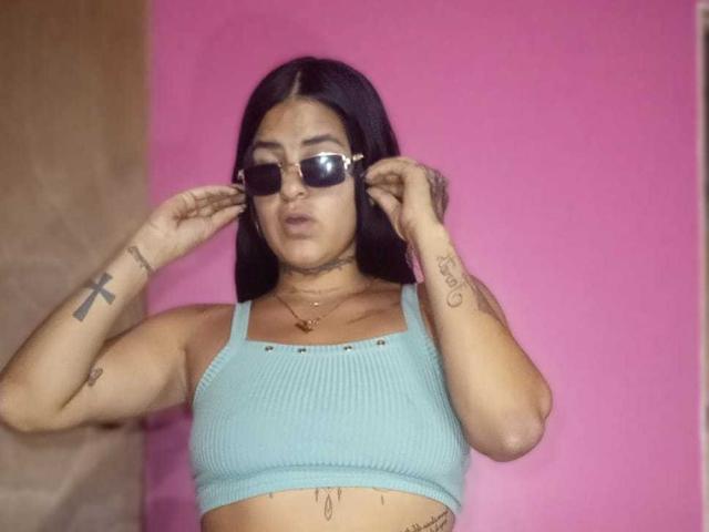 BeautifulTattoo with black hair, wearing sunglasses, a blue ribbed crop top, and tattoos on arms, adjusting her glasses against a pink background.