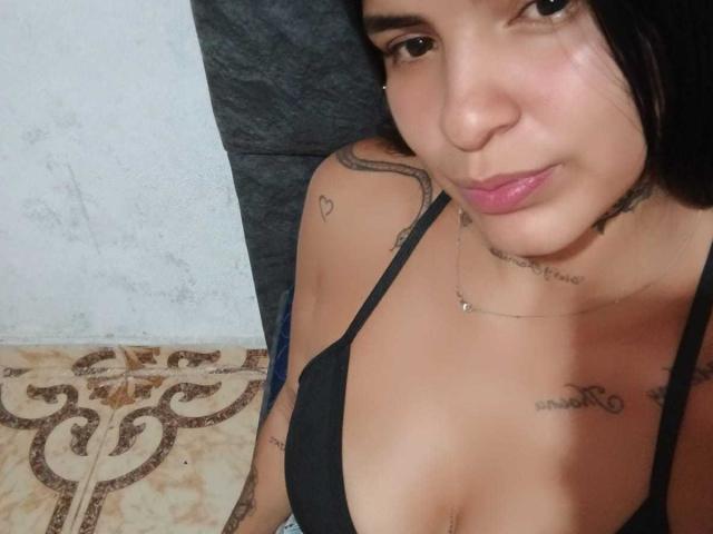 BeautifulTattoo has light brown skin, black hair, and pink lipstick. She's wearing a black top, showing her tits and tattoos on her neck and chest. She's smiling slightly.