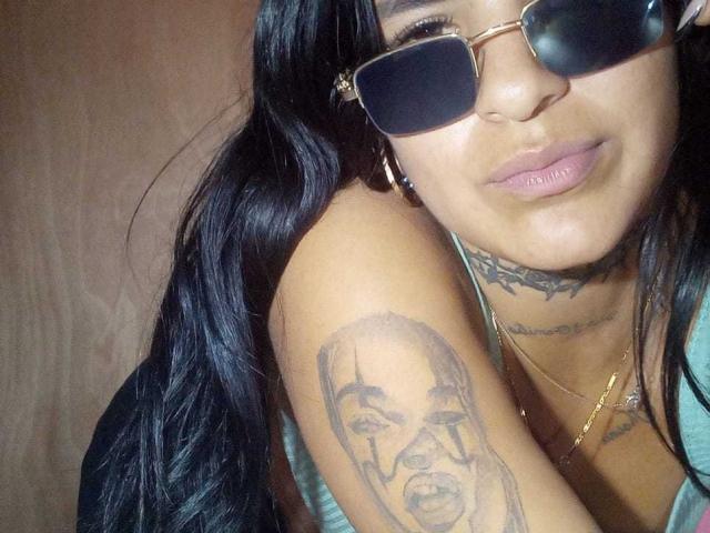 BeautifulTattoo with long black hair, wearing sunglasses, has a detailed tattoo of a bald man on her upper arm, and visible tattoos on her neck, smiling.