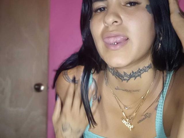 BeautifulTattoo with black hair, blue tank top, gold necklaces, tattoos on neck and chest, sticking out tongue with lip piercing, holding hair. Pink wall, wooden door.