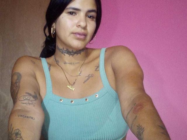 A selfie of BeautifulTattoo, a woman with tan skin and black hair in a ponytail, wearing a teal ribbed tank top. She has multiple tattoos on her arms, chest, and neck, including a star on her collarbone and text on her right arm. She's wearing gold hoop earrings and two necklaces with small pendants. Her expression is neutral. The background is a solid pink color. The image is focused on her upper body.