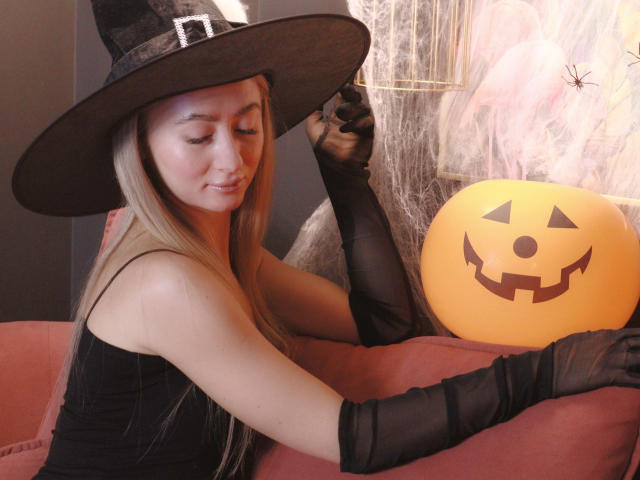 VictoriaLight with long blonde hair, wearing a black witch hat, black sleeveless top, and black gloves, holding a pumpkin with a jack-o'-lantern face, sitting on a pink couch.