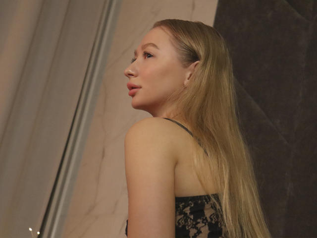 VictoriaLight with long blonde hair, looking over her shoulder, wearing a black lace top, showing her ass, in a dimly lit room, her face turned to the side.