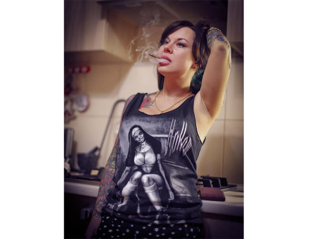 MissTwinkleX stands in a kitchen, smoking a cigarette. She has tattoos on her right arm and left shoulder. She wears a black tank top with a graphic of a woman in a black and white horror image and polka dot pants. She has short dark hair, with one side pulled up, and she's looking at the camera with a slight pout. Smoke curls from her cigarette. The image is brightly lit with a soft focus. Her expression is confident.