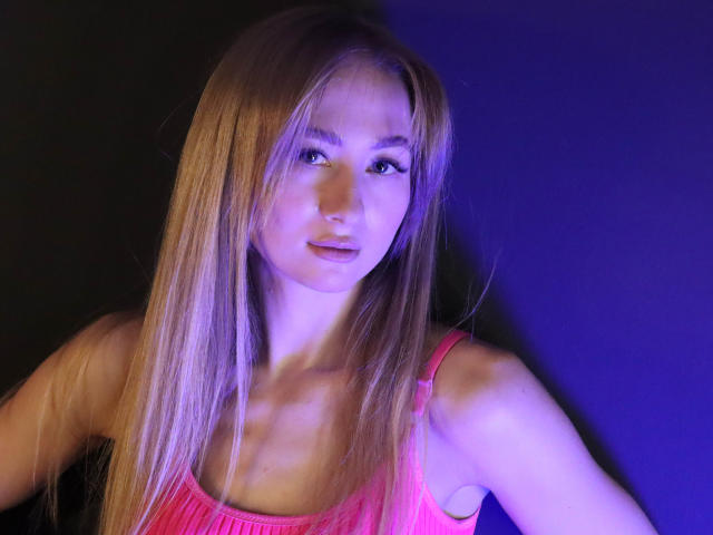 VictoriaLight with long blonde hair, pink tank top, and purple lighting, showing off her tits and a slight smile, looking directly at the camera.