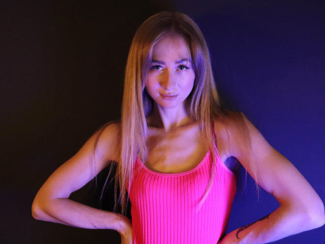VictoriaLight met lang blond haar, een roze geribbelde tanktop, en handen op haar heupen, die haar kleine borsten en een slanke, getoonde billen laat zien, tegen een donkere achtergrond..