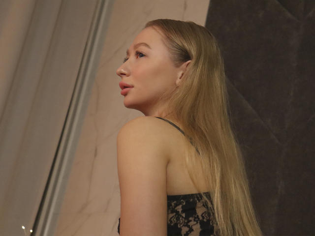 VictoriaLight has long, straight blonde hair. She's turned slightly to the side, showing her profile. She wears a black lace top with thin straps. Her skin is fair, and she has light makeup with pink lips. The background is blurred, with beige and dark brown textures. She has small, subtle earrings. The image focuses on her upper body and face.