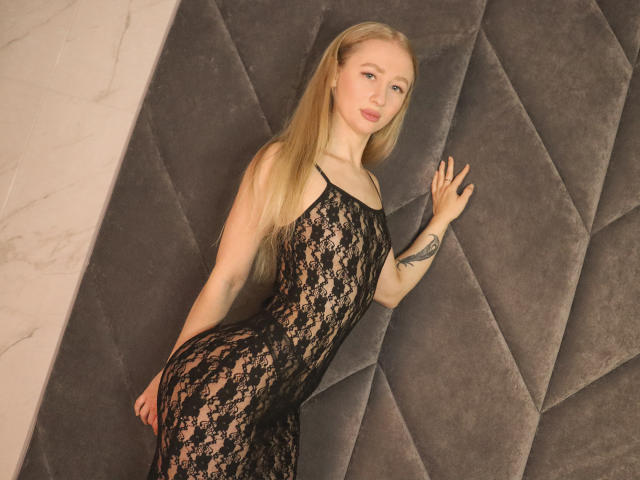 Victoria, vestita con un abito nero in pizzo, che mette in mostra glutei e seno, appoggiata a una parete grigia imbottita, con lunghi capelli biondi e un tatuaggio sul braccio destro..