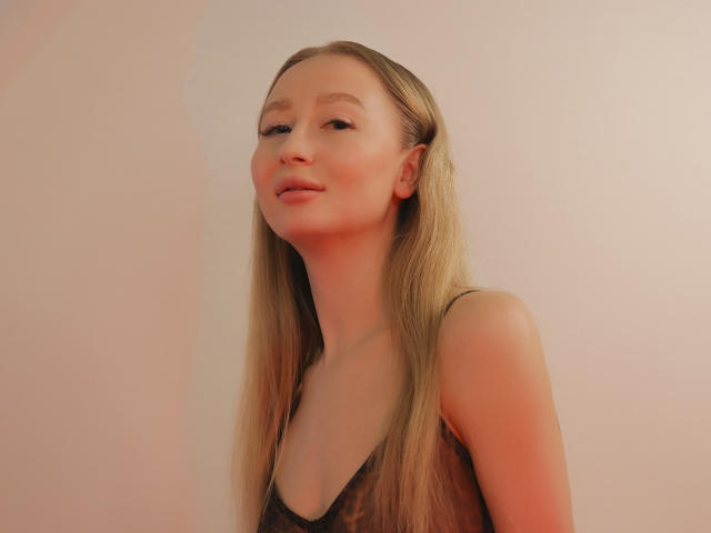 Portrait of VictoriaLight with straight, long blonde hair. She has light skin and a subtle smile, looking slightly upward. She's wearing a black, sheer, spaghetti-strap top. Soft, warm lighting casts a reddish-orange hue on her face. The background is a plain, light beige wall. Her hair is centered, and her top's texture is slightly transparent. The image has a clean, minimalist composition.