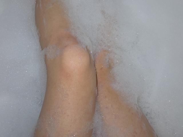 Le gambe di Amberia sono parzialmente immerse in acqua schiumosa, con le ginocchia leggermente piegate e le dita dei piedi visibili, che mostrano una pelle liscia e chiara..