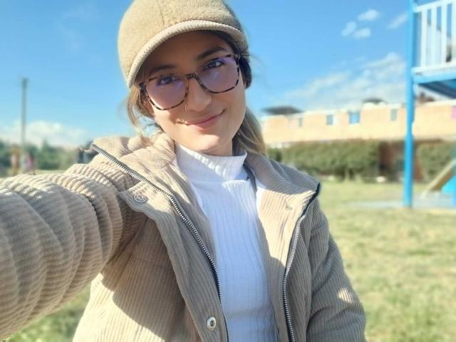 Kaiix with glasses, beige beanie, ribbed tan jacket, white turtleneck, smiling, taking selfie, blue sky, green grass, and blue playground in background. Kaiix's tits are small.