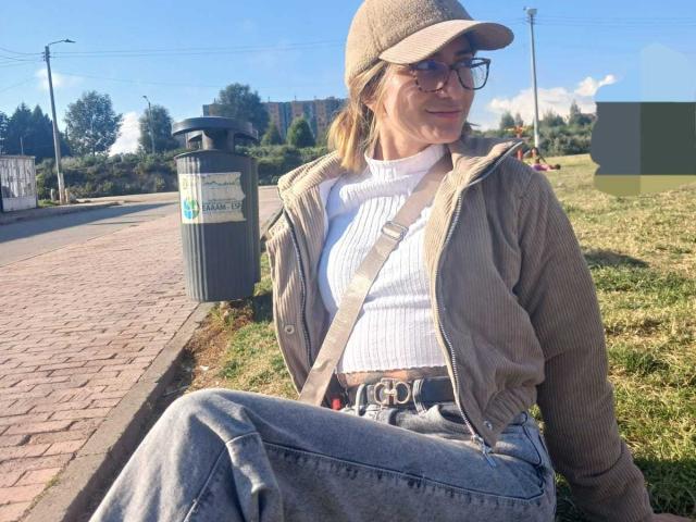 Kaiix with a cap, glasses, white ribbed top, brown jacket, and grey jeans, sitting on grass, holding trash can, sunny day, looking happy, belt with buckle.