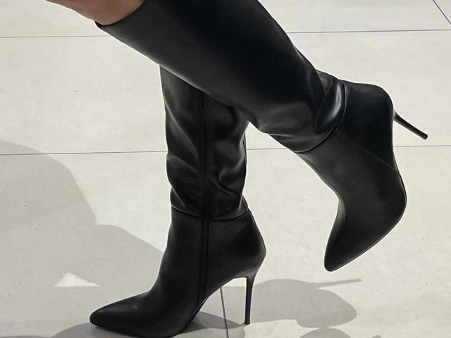 DeesseBleue's black, shiny, high-heeled, thigh-length boots with pointed toes and stiletto heels, showing one boot slightly raised, showcasing the sleek, glossy texture.