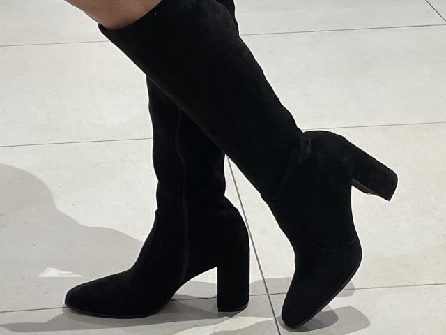 DeesseBleue's black, high-heeled, suede boots with a blocky, square heel are standing on a white-tiled floor. Her legs are crossed at the ankles.
