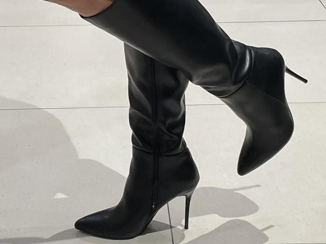 DeesseBleue's black, high-heeled, knee-boots with a sleek, shiny leather texture and pointed toes are stepping on a light gray, tiled floor, showing a slight shadow.