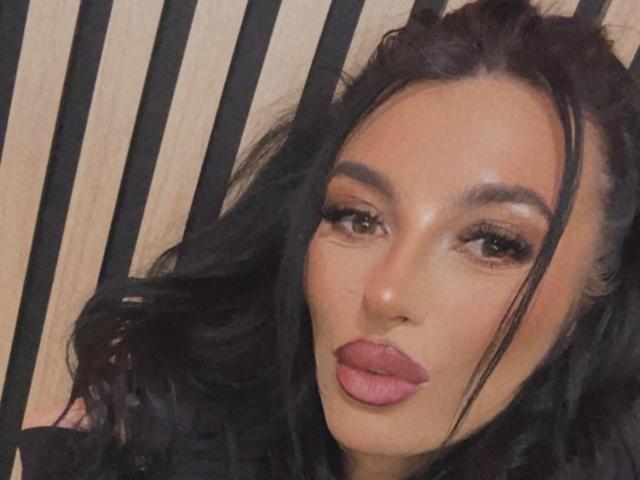 KinkyAmelie has glossy brown eyes, full pink lips, and wavy black hair with a slight part, looking directly at the camera with a slightly pouty expression.