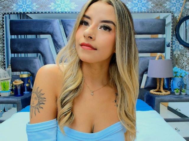 CloeRouss with blonde hair, blue off-shoulder top, small tits, sun tattoo on right shoulder, red lipstick, sitting on bed, blue and gray background, looking at camera.