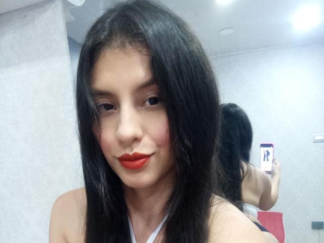 MadissonFontaine with long black hair, red lipstick, and a slight smile, holding a phone in a mirror selfie, showing her bare shoulders and a reflection of her ass.