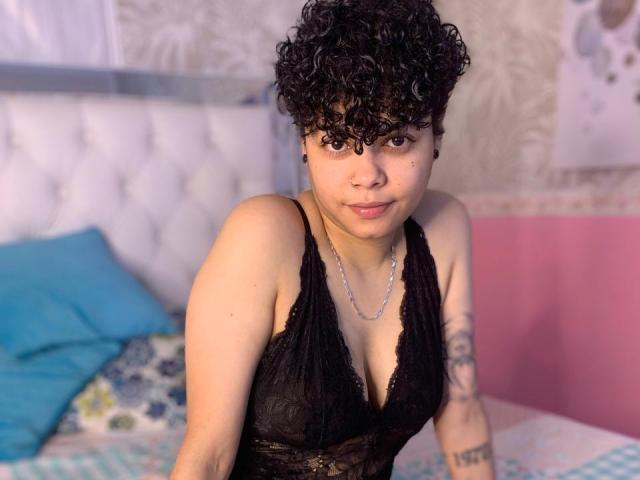 SashaVenom with short curly black hair, wearing a black lace halter top, showing her small tits and a tattoo on her right arm, sitting on a bed.