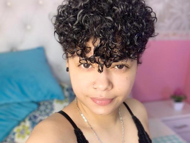 Close-up of SashaVenom, a young person with curly black hair, light brown skin, and a small nose ring. SashaVenom wears a black lace top and a silver necklace. Their brown eyes are centered, and they have a slight, neutral expression. The background is blurred, featuring blue and pink pillows. The texture of SashaVenom's hair is tight and curly, contrasting with the smoothness of their skin. The image has a soft, natural light.