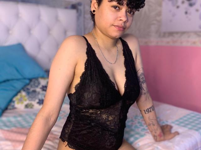 SashaVenom, a curvy woman with short curly black hair, wearing a black lace lingerie top, showing small tattoos on her right arm, sitting on a bed.